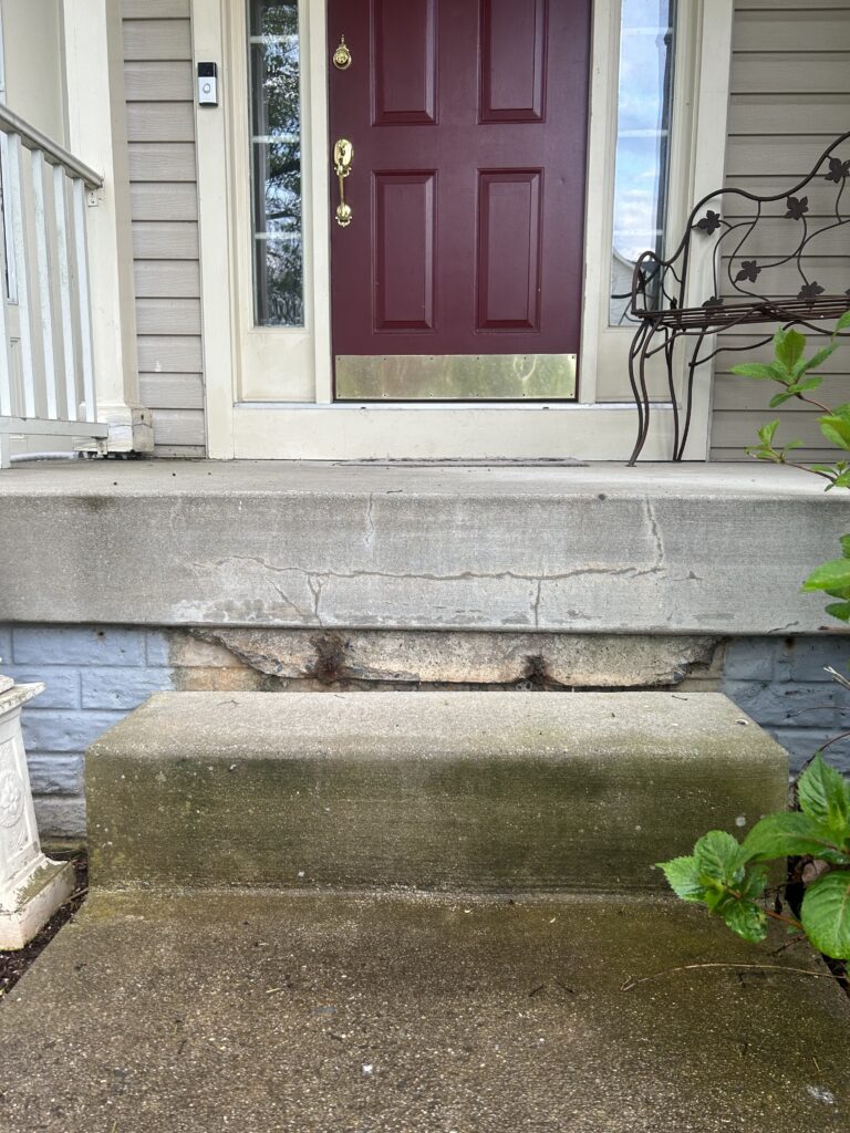Photo Apr 15 2024 7 33 34 AM - Poly Concrete Jacking Front entry with a burgundy door, brass hardware, and a brass kick plate on a concrete porch with cracked steps.