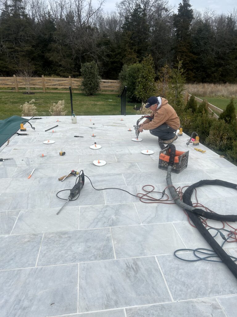 Photo Dec 07 2023 10 57 27 AM - Poly Concrete Jacking Worker kneeling on a tiled patio, using a power tool to install or adjust tiles with scattered tools around him in a backyard setting.