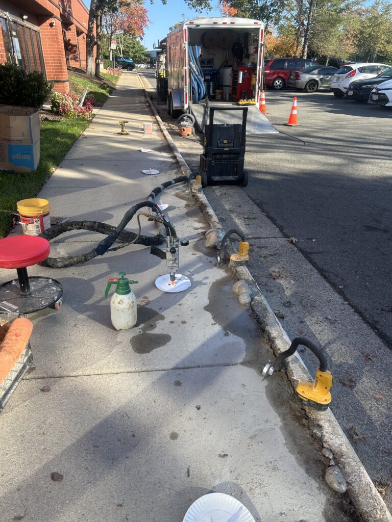 Photo Oct 17 2024 9 43 52 AM - Poly Concrete Jacking Construction work on sidewalk: utility trailer with hoses and tools, orange cones, and exposed trench along curb with equipment nearby